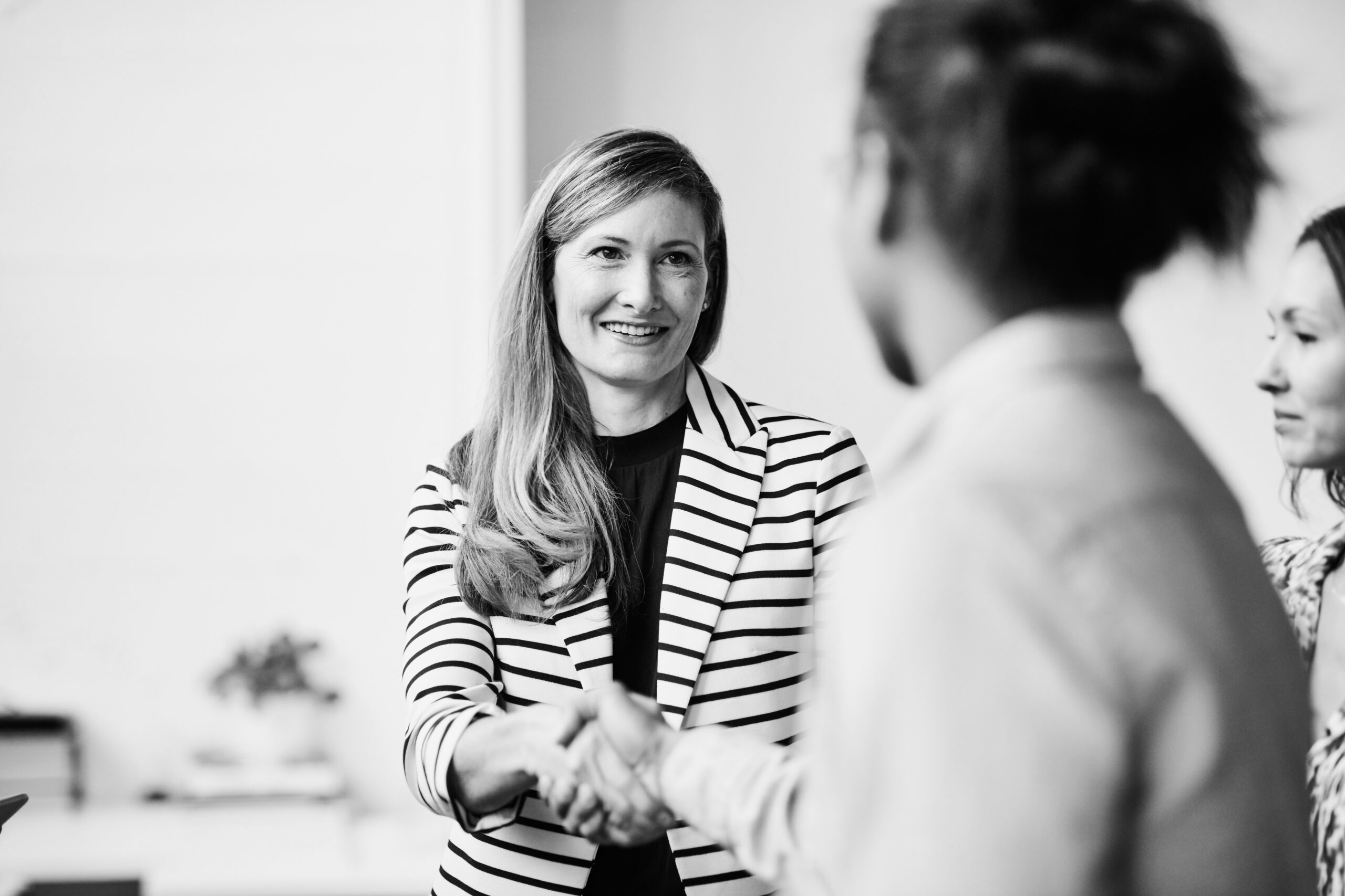 A woman in a striped blazer shakes hands with another person, while a third person stands nearby, all appearing to be engaged in a professional setting.