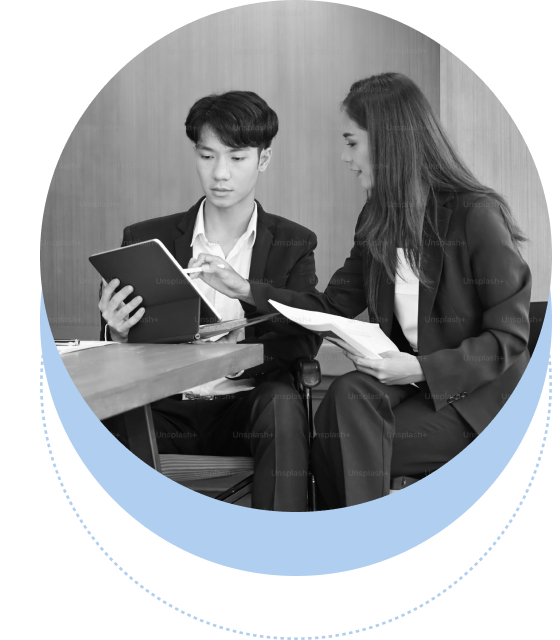 Two people in business attire are sitting at a table. One is holding a tablet and the other is gesturing towards it while holding documents. They appear to be discussing work.