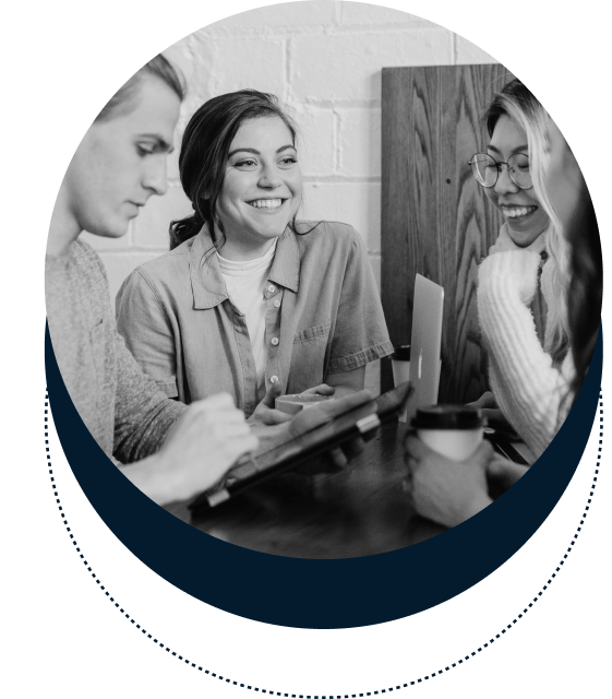 Three people sit around a table, engaging in conversation. One person holds a tablet, another a laptop, and a third holds a cup. They are smiling and seem to be talking collaboratively.
