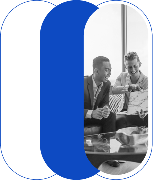 Two men in business attire sit in an office, looking at a document together and smiling.