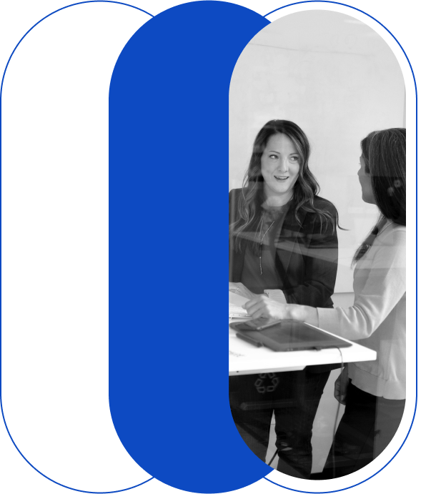 Two women are standing and conversing at a table with a laptop on it, inside a modern office space. A graphic design element frames the image.