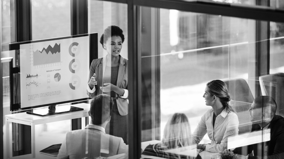 A businesswoman presents charts and graphs on a screen to a group of colleagues in a modern office meeting room.