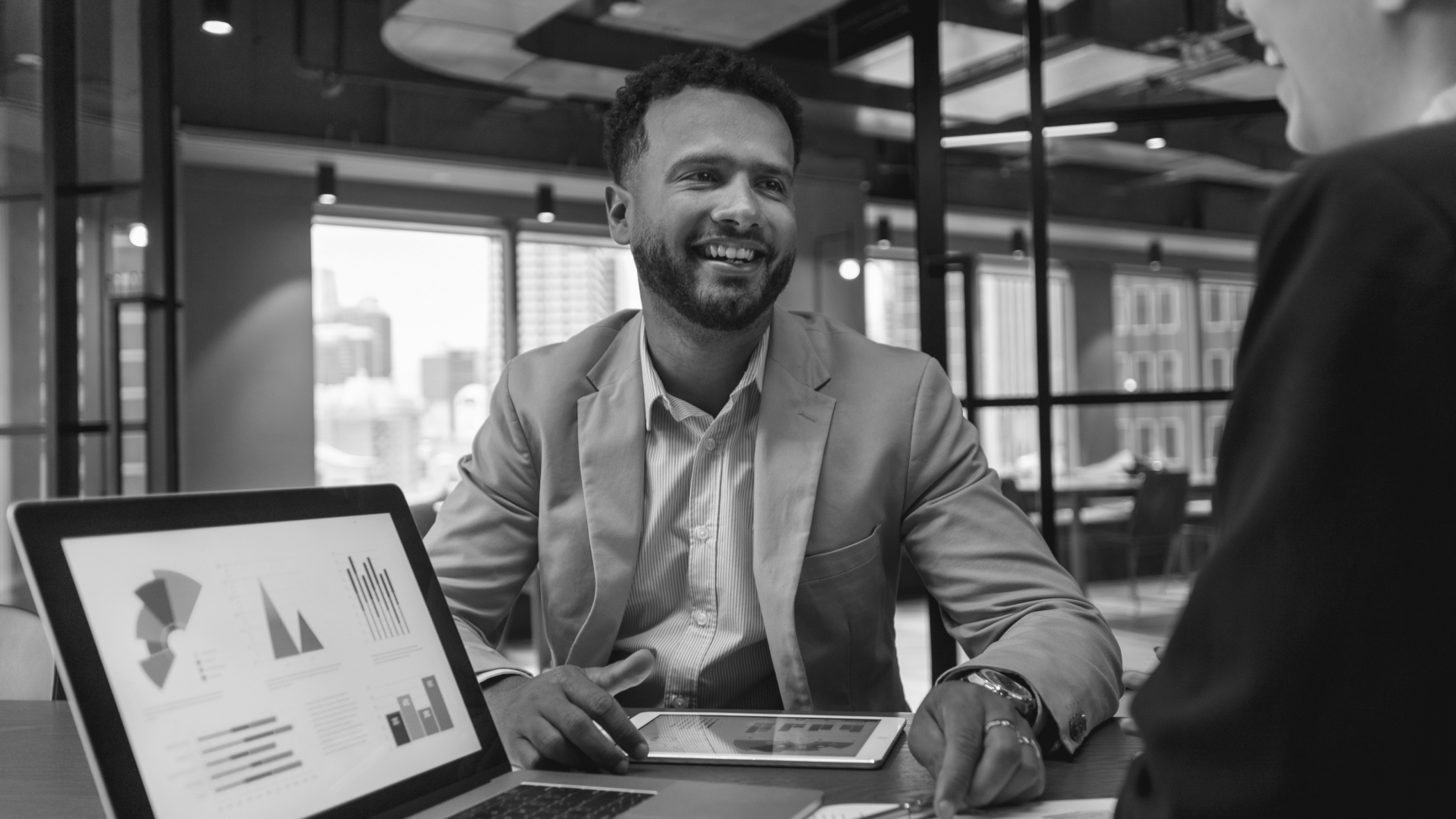 A man in a suit smiles during a business meeting in an office, with charts displayed on a laptop and paper.