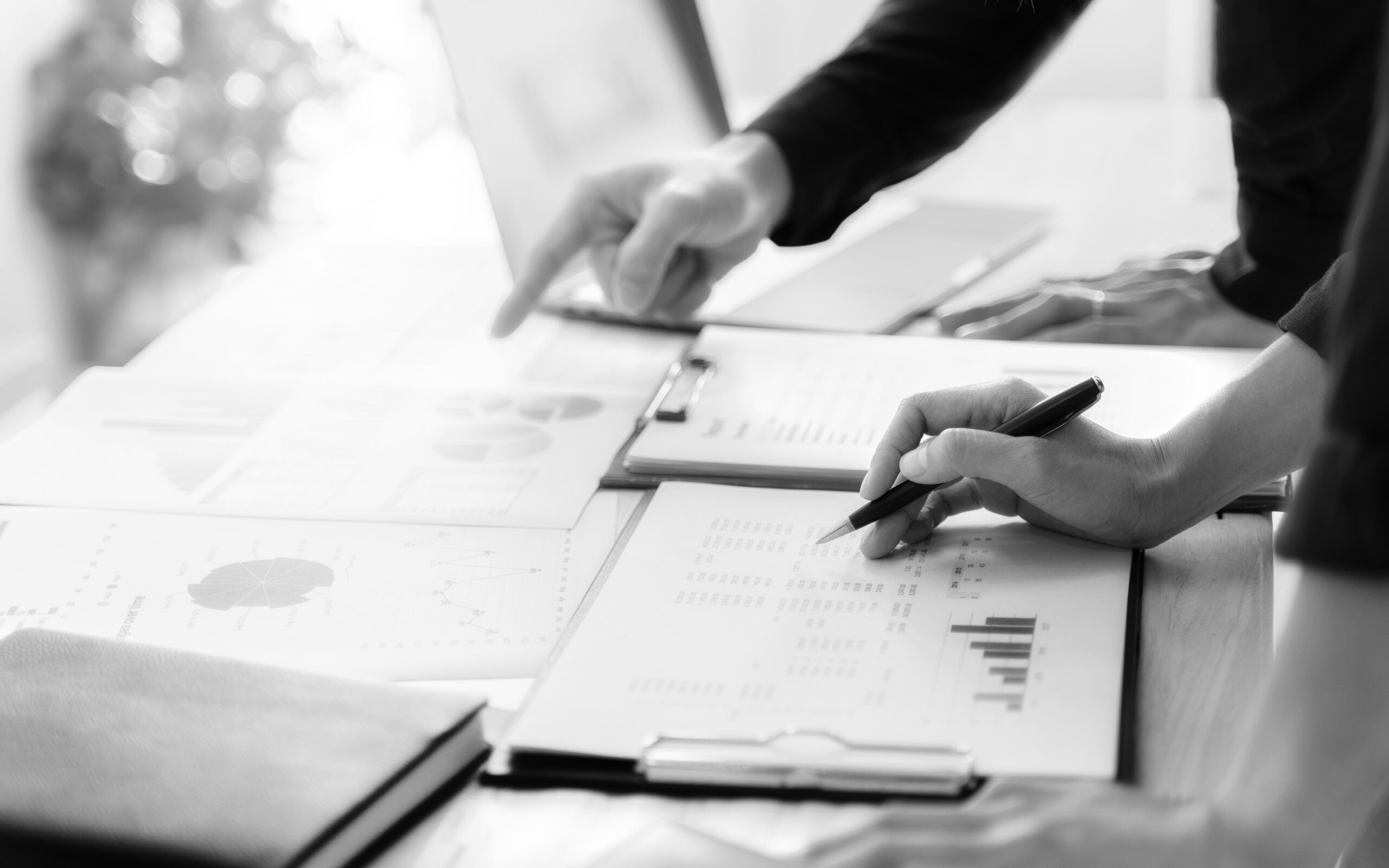 Two people reviewing charts and graphs on paper at a desk. One person points, while the other writes with a pen. Documents include bar graphs and pie charts.